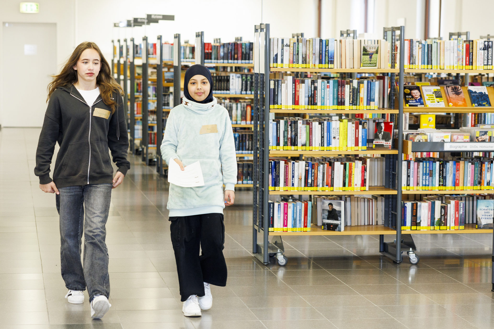 Zukunftstag in der Stadtbibliothek. Zwei Mädchen gehen an Bücherregalen vorbei. (Wird bei Klick vergrößert)